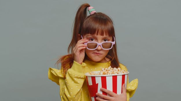 Excited young toddler school girl in 3D glasses eating popcorn, watching interesting tv serial, sport game, film, online social media movie content. Teen female child kid on studio gray background