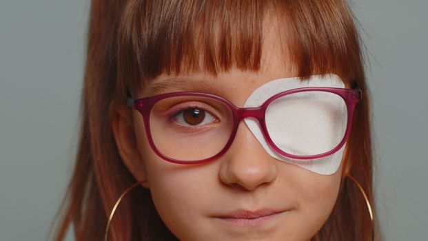 Close-up portrait sick ill preteen girl having protective bandage plaster patch scratch on one eye. Injury, trauma, blind. Child kid wearing glasses at gray background. Medical eyesight vision therapy