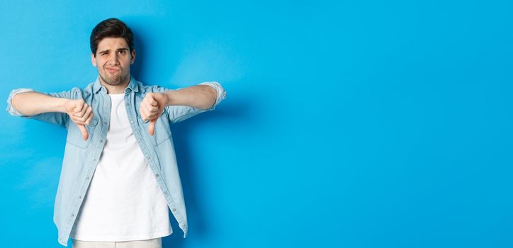 Skeptical and disappointed 25 years old man in casual outfit dislike something bad, showing thumbs-down and grimacing displeased, standing against blue background.