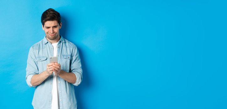 Disappointed and skeptical guy looking at phone, standing over blue background.