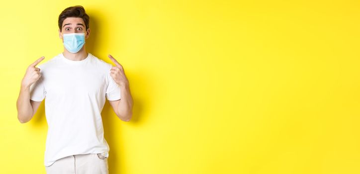 Concept of coronavirus, pandemic and social distancing. Young surprised man pointing at medical mask on face, yellow background. Copy space