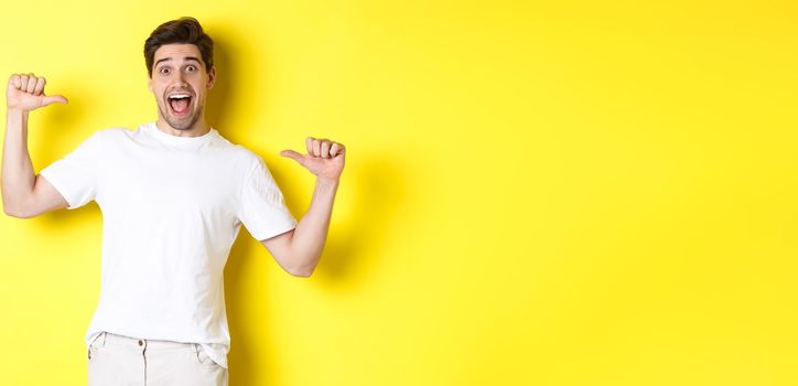 Excited man looking happy, pointing at himself with amazement, standing over yellow background.
