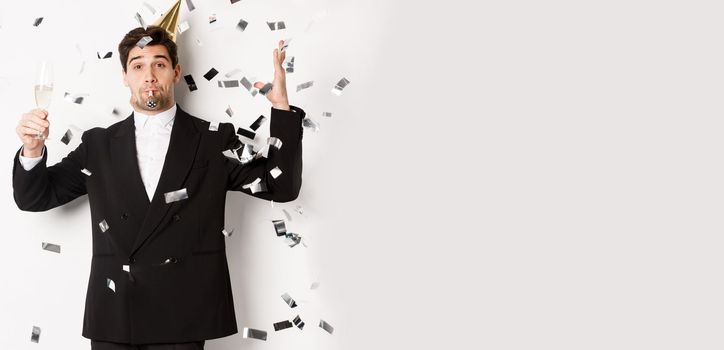 Handsome party guy in black suit having fun, celebrating new year, blowing whistle and drinking champagne while confetti falling, wishing happy holidays, standing against white background.