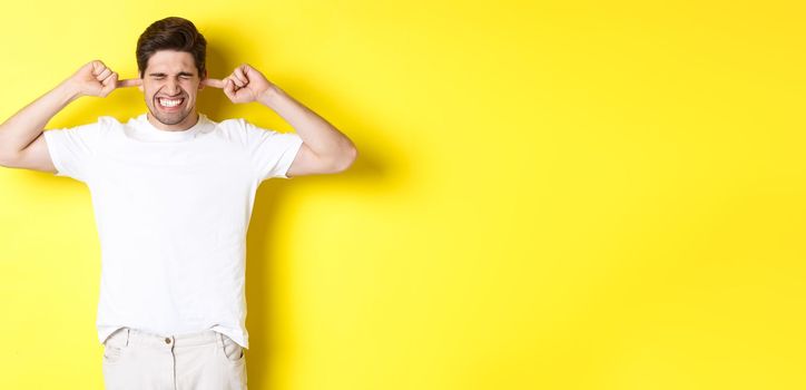 Annoyed man grimacing and shutting ears, complaining on loud noise, standing against yellow background. Copy space