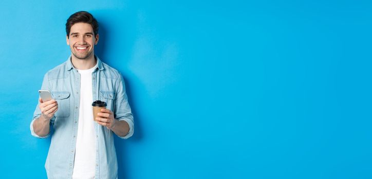 Young modern man drinking coffee and using mobile phone, smiling at camera, standing over blue background.