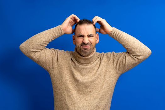 Doubtful bearded latin man thinking, scratching his head and trying to find a solution, isolated over blue background.