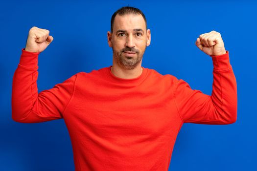 Hispanic male happy unshaven strong sporty fitness student man 40s in red trendy sweater showing biceps muscles on hand isolated on blue color background studio portrait