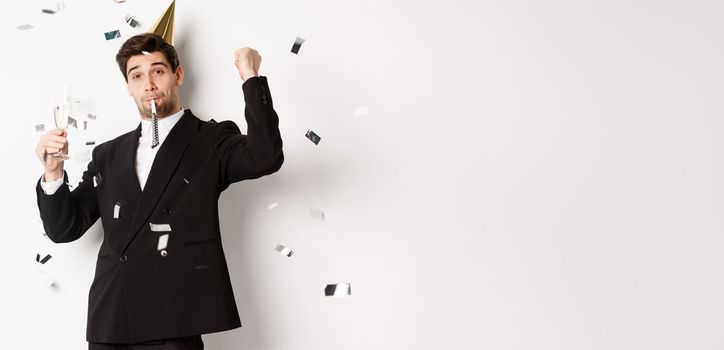 Handsome party guy in black suit having fun, celebrating new year, blowing whistle and drinking champagne while confetti falling, standing happy against white background.