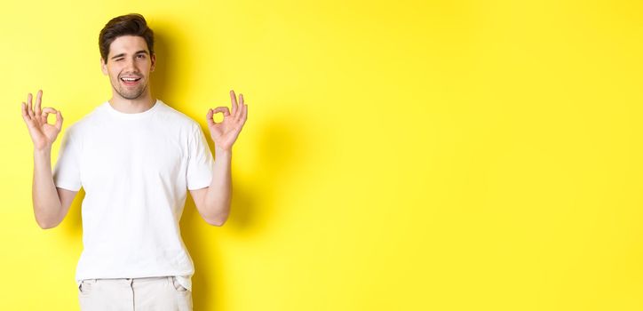 Confident handsome man winking, showing okay signs in approval, like something good, standing over yellow background.