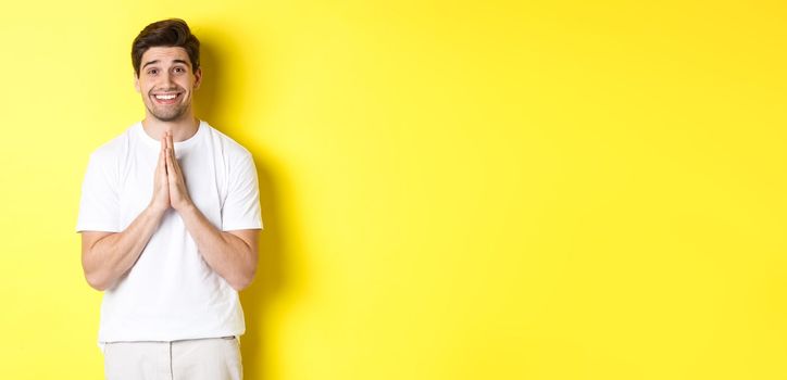 Hopeful man asking for help, begging favour, need something and smiling, standing over yellow background.