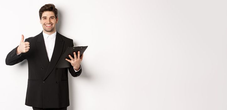 Portrait of confident businessman in black suit, holding clipboard with documents and showing thumb-up in approval, praise good job, standing against white background.