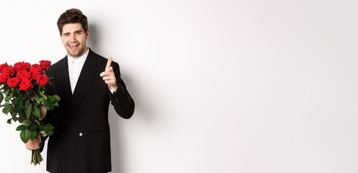 Image of handsome romantic guy in black suit, holding bouquet of roses and pointing at camera, congratulating with holiday, standing against white background.