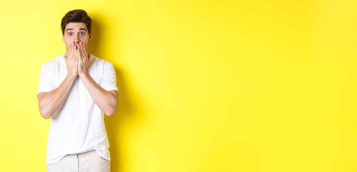 Handsome man looking shocked and speechless, holding hands on mouth, standing over yellow background.