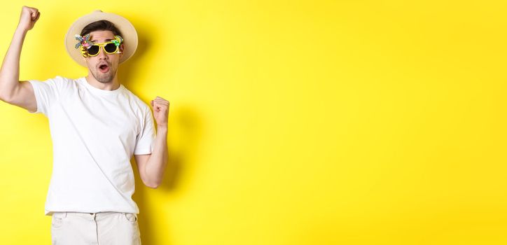 Concept of tourism and lifestyle. Happy guy tourist enjoying trip, rooting for you, fist pump and triumphing, going on journey in summer hat and sunglasses, yellow background.