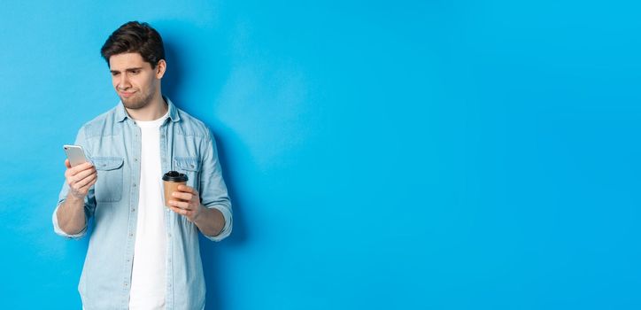 Skeptical and disappointed guy reading upsetting message on phone, holding coffee cup, standing over blue background.