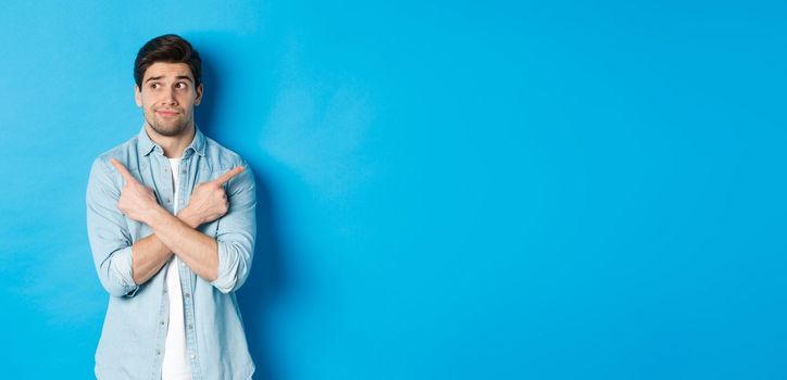 Portrait of indecisive adult man pointing fingers sideways but looking left, making choice between two products, standing against blue background.