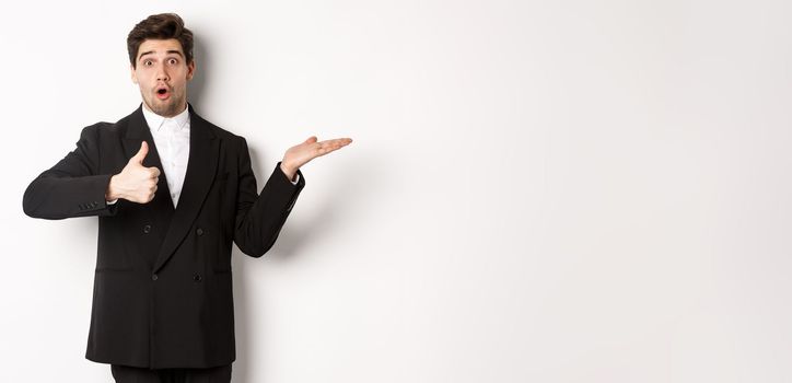 Portrait of hadnsome bearded man in formal suit, showing thumb-up and holding product in hand over white copy space, recommending product, standing over white background.