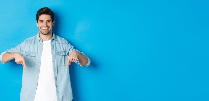 Handsome smiling adult man introduce product, pointing fingers down at promotion, standing against blue background.