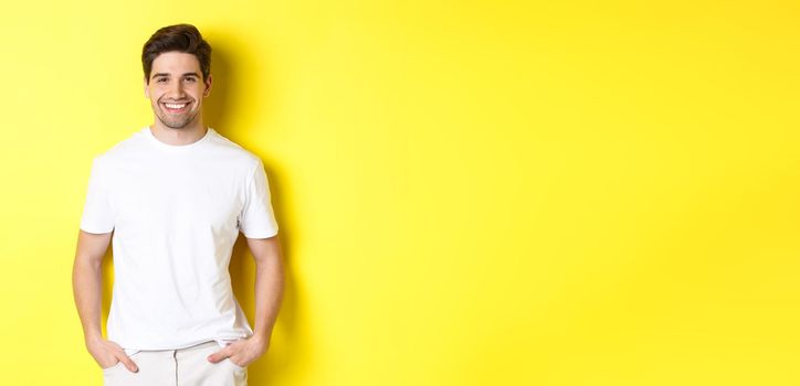 Young handsome man smiling at camera, holding hands in pockets, standing against yellow background.