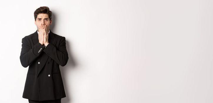 Portrait of shocked handsome businessman in suit, reacting to terrible situation, gasping and covering mouth with hands, standing startled against white background.