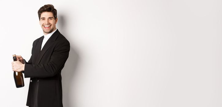 Handsome smiling man in trendy suit open a bottle of champagne, celebrating holidays, standing over white background.