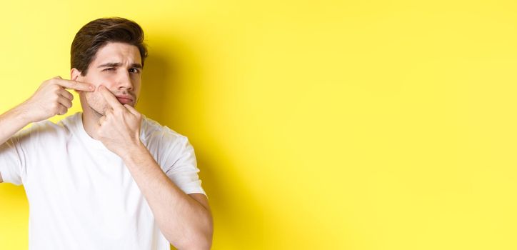 Young man pop a pimple on cheek, standing over yellow background. Concept of skin care and acne.