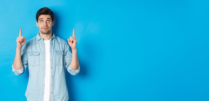 Portrait of displeased and skeptical male model pointing fingers up, looking at something unpleasant, standing against blue background.