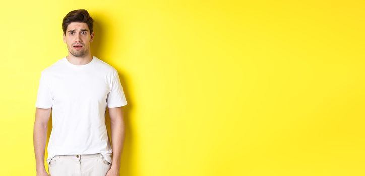 Image of confused and nervous man looking at something strange, frowning anxious, standing against yellow background.