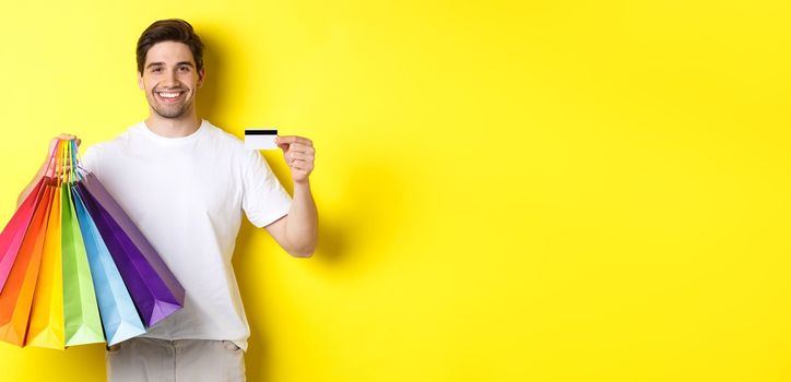 Young man shopping for holidays, holding paper bags and recommending bank credit card, standing over yellow background.