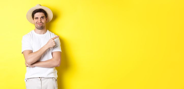 Skeptical male tourist complaining, pointing finger right at something bad or lame, standing unamused against yellow background.