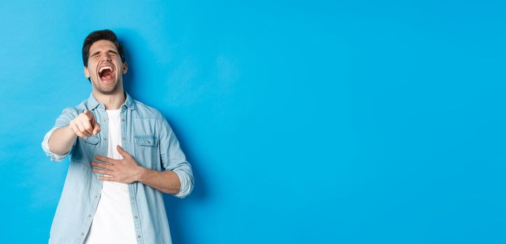 Adult man in casual outfit laughing out loud and pointing at you, looking at something funny, chuckling while standing against blue background.