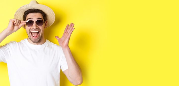 Concept of tourism and holidays. Close-up of surprised man shouting for joy, enjoying vacation, wearing sunglasses with summer hat, yellow background.