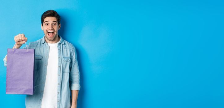 Concept of shopping, holidays and lifestyle. Excited handsome guy holding paper bag with gift and looking happy, standing over blue background.