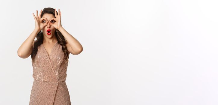 Christmas party and celebration concept. Woman in glamour dress and red lips looking at something amazing, staring through hand binoculars, standing over white background.