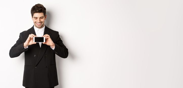 Portrait of handsome bearded man in black suit, showing you smartphone screen and smiling pleased, standing against white background.