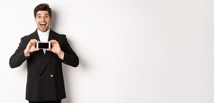 Image of cheerful, handsome man in black suit, showing smarthone screen and looking amazed, standing against white background.