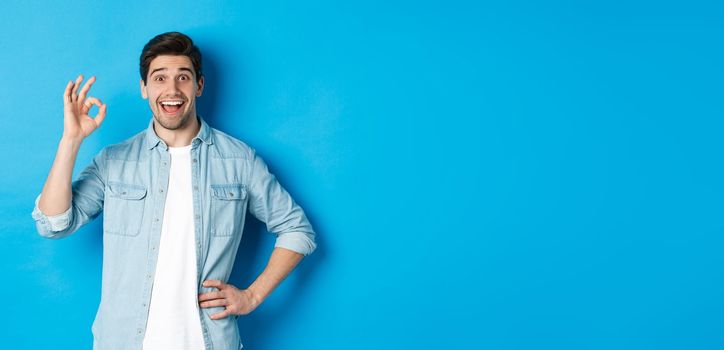 Smiling happy man showing ok sign and looking pleased, approving something good, standing against blue background.