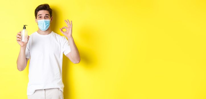 Concept of covid-19, quarantine and lifestyle. Satisfied young man in medical mask showing good hand sanitizer, make okay sign and recommending antiseptic, yellow background.