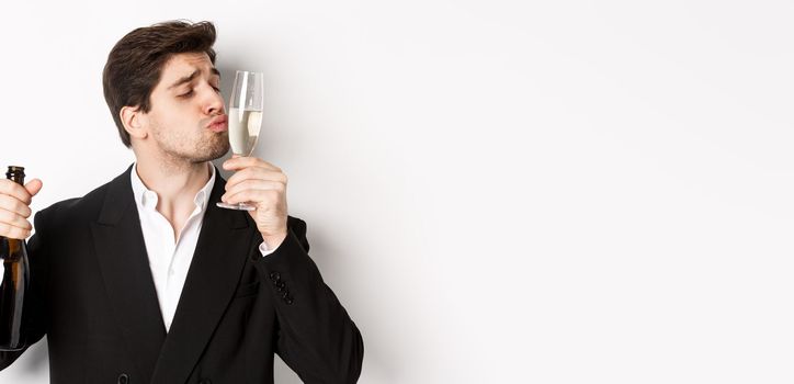 Close-up of handsome man in suit, kissing glass with champagne, getting drunk on a party, standing against white background.