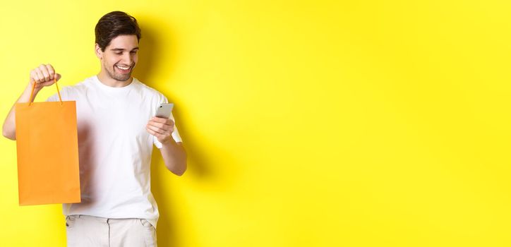 Concept of discounts, online banking and cashback. Happy guy showing shopping bag and looking satisfied at mobile screen, yellow background.