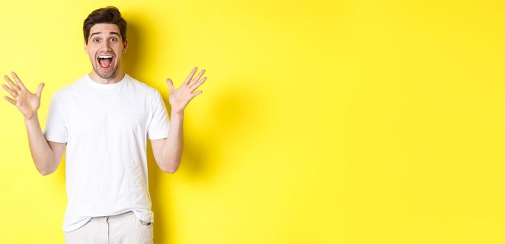 Portrait of excited and surprised man reacting to big promo offer, shouting for joy and triumphing, standing over yellow background.