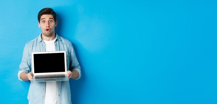 Man showing advertisement on laptop screen and looking amazed, saying wow and looking at camera, standing against blue background.