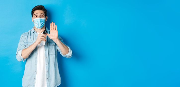 Concept of covid-19, pandemic and social distancing. Cheerful guy in medical mask showing how to disinfect hands with sanitizer, using antiseptic, preventing virus spread, blue background.