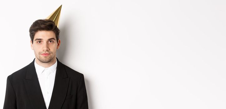 Close-up of handsome man in party hat and trendy suit, celebrating new year, standing against white background.