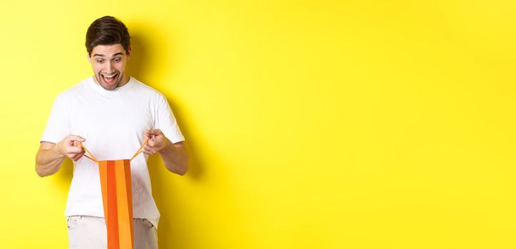 Excited guy open bag with gift, looking inside with amazement and happy face, standing against yellow background.