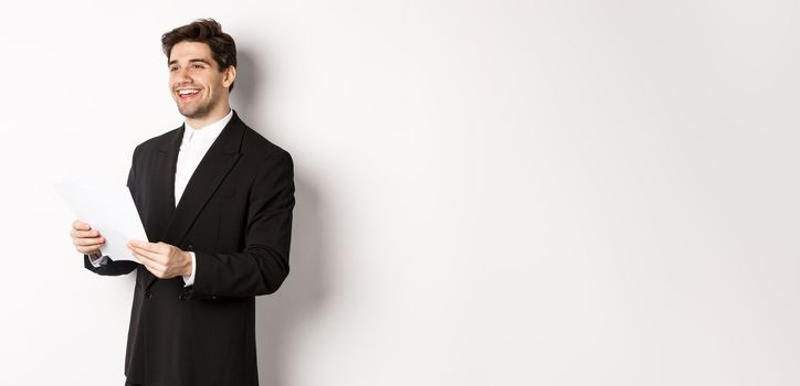 Image of handsome businessman in suit, holding documents and smiling, standing against white background.