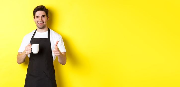 Handsome barista in black apron holding coffee cup, pointing finger at you, inviting visit cafe, standing over yellow background.
