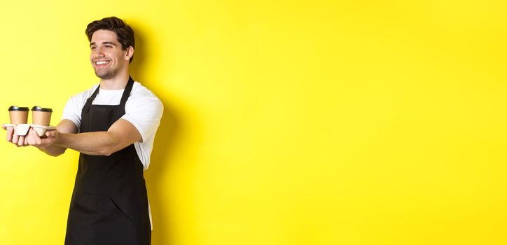 Friendly barista in black apron giving takeaway order, holding two cups of coffee and smiling, standing over yellow background.