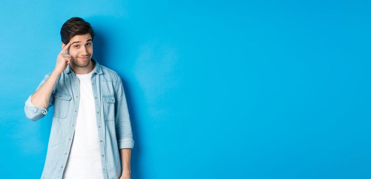 Young thoughtful man pointing at head, asking to think about it, giving a hint, standing in casual clothes over blue background.