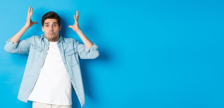 Distressed guy showing mind blowing gesture, looking frustrated and anxious, standing against blue background.